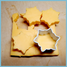 Un moule, de style coupe biscuit, en forme de flocon appuyé sur une tranche de fromage à côté de morceaux de fromages en forme de flocon. 