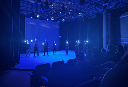 Photo de la scène d’un théâtre et d’une partie des spectateurs, la salle est dans une semi-obscurité bleutée. Les acteurs qui sont sur scène ont une lampe de poche allumée vers le public. 