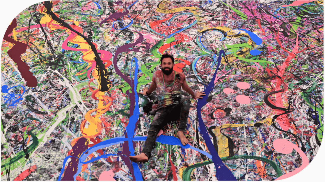 Photo de l’artiste Sacha Jafri assis sur sa gigantesque toile abstraite très colorée. 