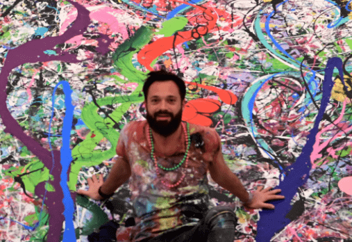 Photo de l’artiste Sacha Jafri assis sur sa gigantesque toile abstraite très colorée. 
