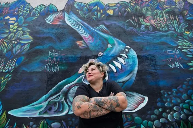 Une photo de l’artiste graffiti franco-ontarienne Mique Michelle. Elle a les bras croisés devant un graffiti de baleine. 