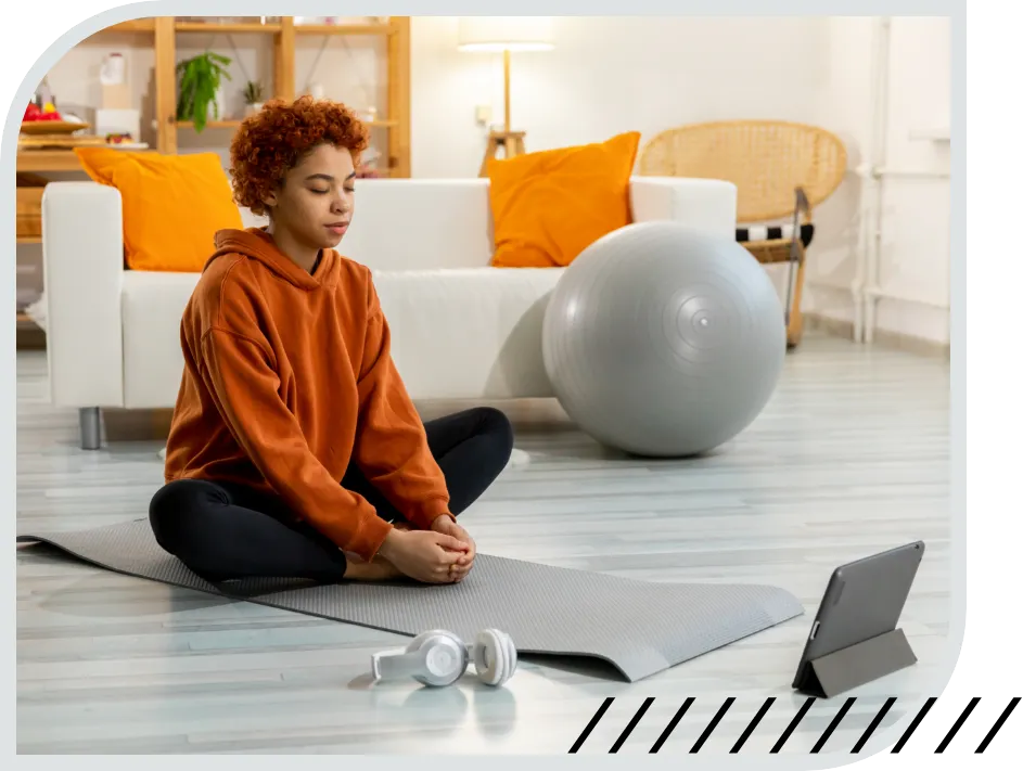 Une personne assise en tailleur les yeux fermés sur un tapis de yoga. Une tablette électronique lui fait face.