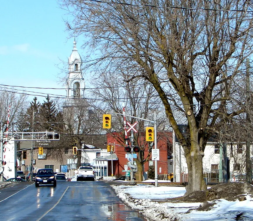 Photo d’un quartier de Casselman. Le clocher de l’église point en arrière-plan. Un carrefour de deux rues avec feux de circulation figure à l’avant-plan avec quelques bâtiments entre les deux. La voie ferrée traverse l’une des rues.