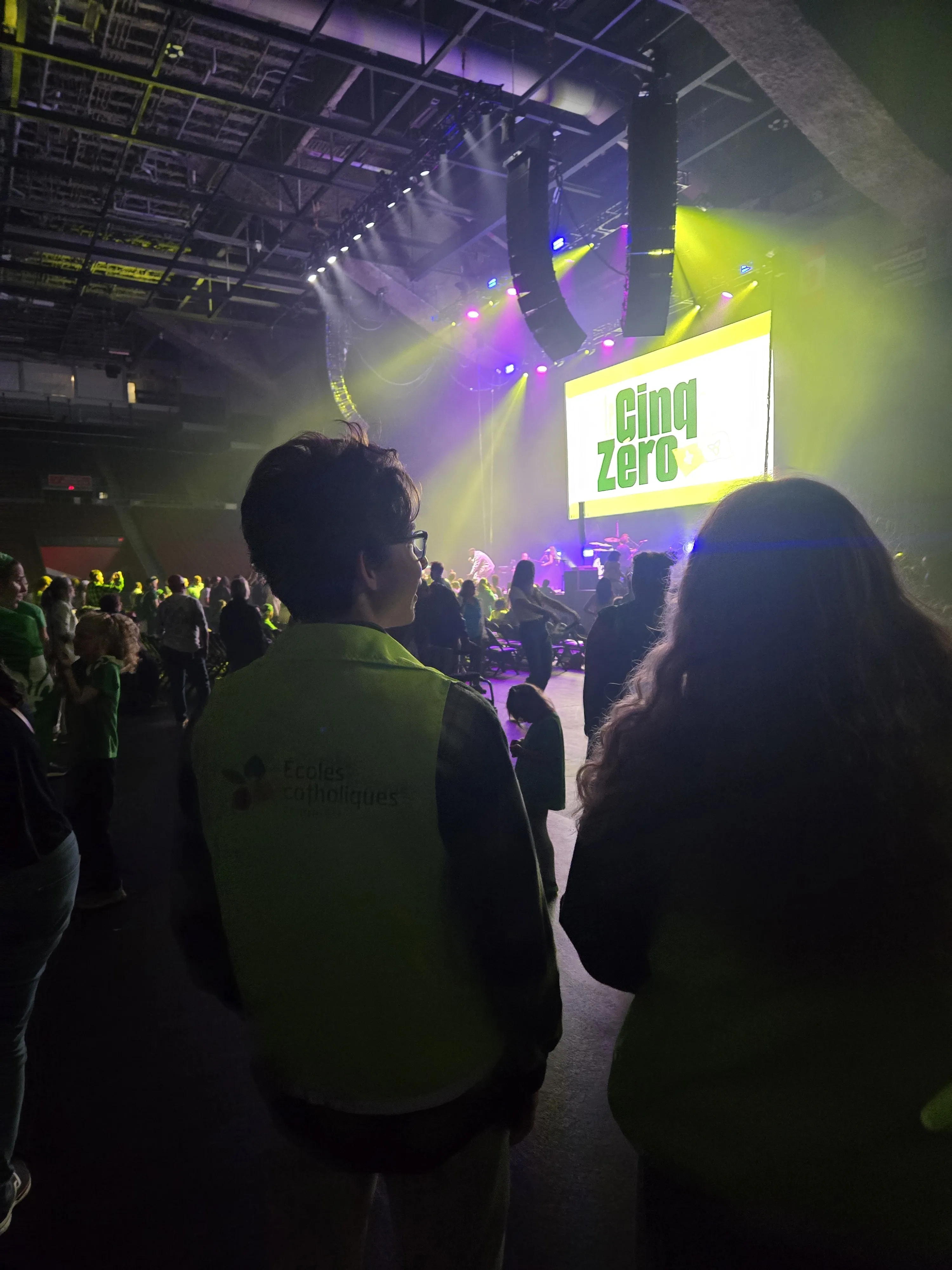 Photo de l’événement Cinq Zéro avec Philippe Beauchemin et Hailey Mainville. À l’avant, un écran lumineux. Une personne au premier plan porte un dossard clair avec le logo des écoles catholiques. Des techniciens, du public et des bénévoles sont visibles.