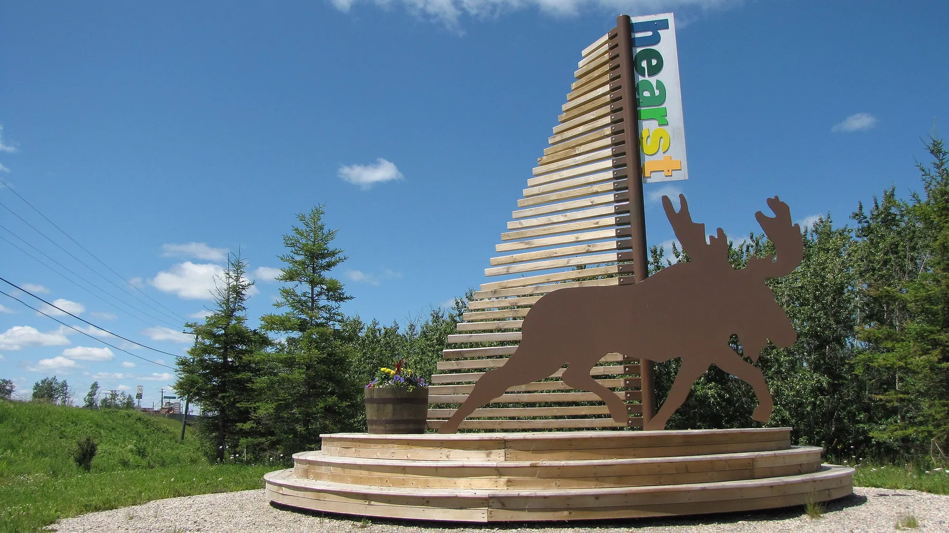 Structure en bois avec silhouette d’orignal et panneau coloré « Hearst », sous un ciel bleu, entourée de verdure et d’arbres, symbole d’accueil et nature.