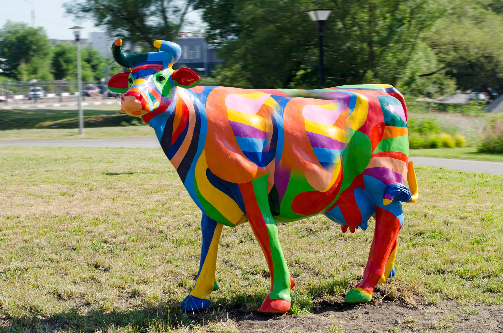 Une statue de vache grandeur nature ornée de motifs géométriques et de rayures multicolores, posée sur un terrain gazonné.