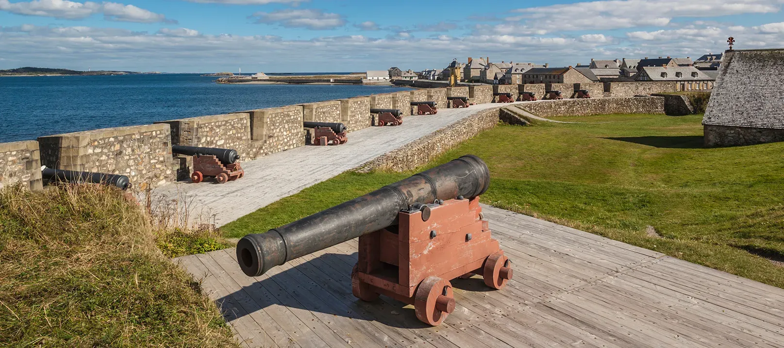 Photo d’un canon dans la forteresse de Louisbourg. Derrière, une muraille de pierre longe la mer. D’autres canons sont alignés le long des remparts.