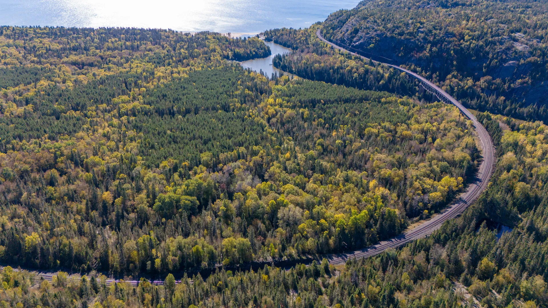 Vue aérienne d’une autoroute sinueuse traversant une vaste et dense forêt et une petite rivière, puis contournant ensuite un flanc de montagne tout en longeant une plus grande étendue d’eau (lac ou fleuve) visible à l’arrière-plan.