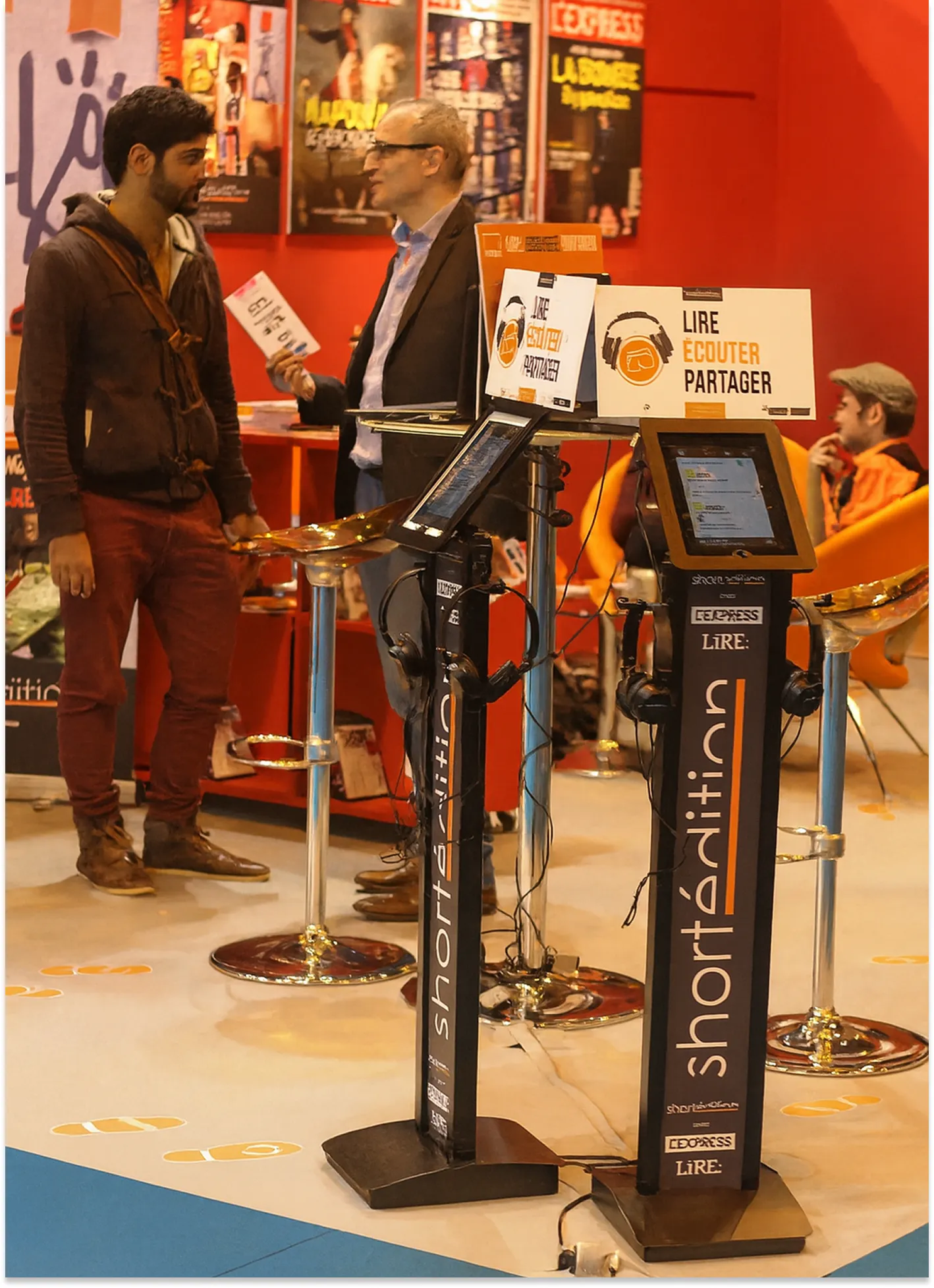 Dans un espace public ouvert, deux hommes discutent derrière des bornes Shortédition, semblables à des lutrins avec tablette électronique. Une affiche au-dessus, illustrant des écouteurs, porte le slogan : « Lire, écouter, partager ».