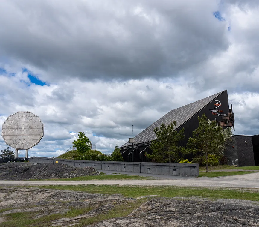 Photo de la sculpture du Gros cinq sous (Big Nickel) dans le Grand Sudbury. La sculpture est dans un terrain vague à côté d’un édifice sur la façade duquel on peut lire « Dynamic Eart – Terre dynamique ».