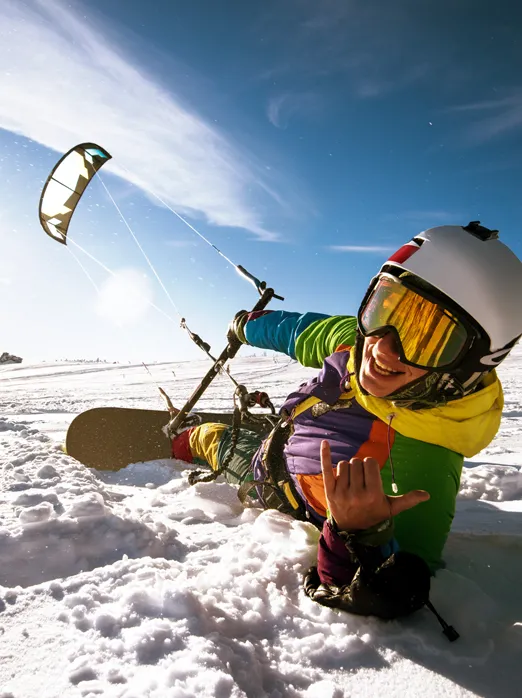 Photo d’une personne souriante qui pratique le ski cerf-volant. La planche à neige aux pieds, elle est allongée sur la neige et tient un cerf-volant.