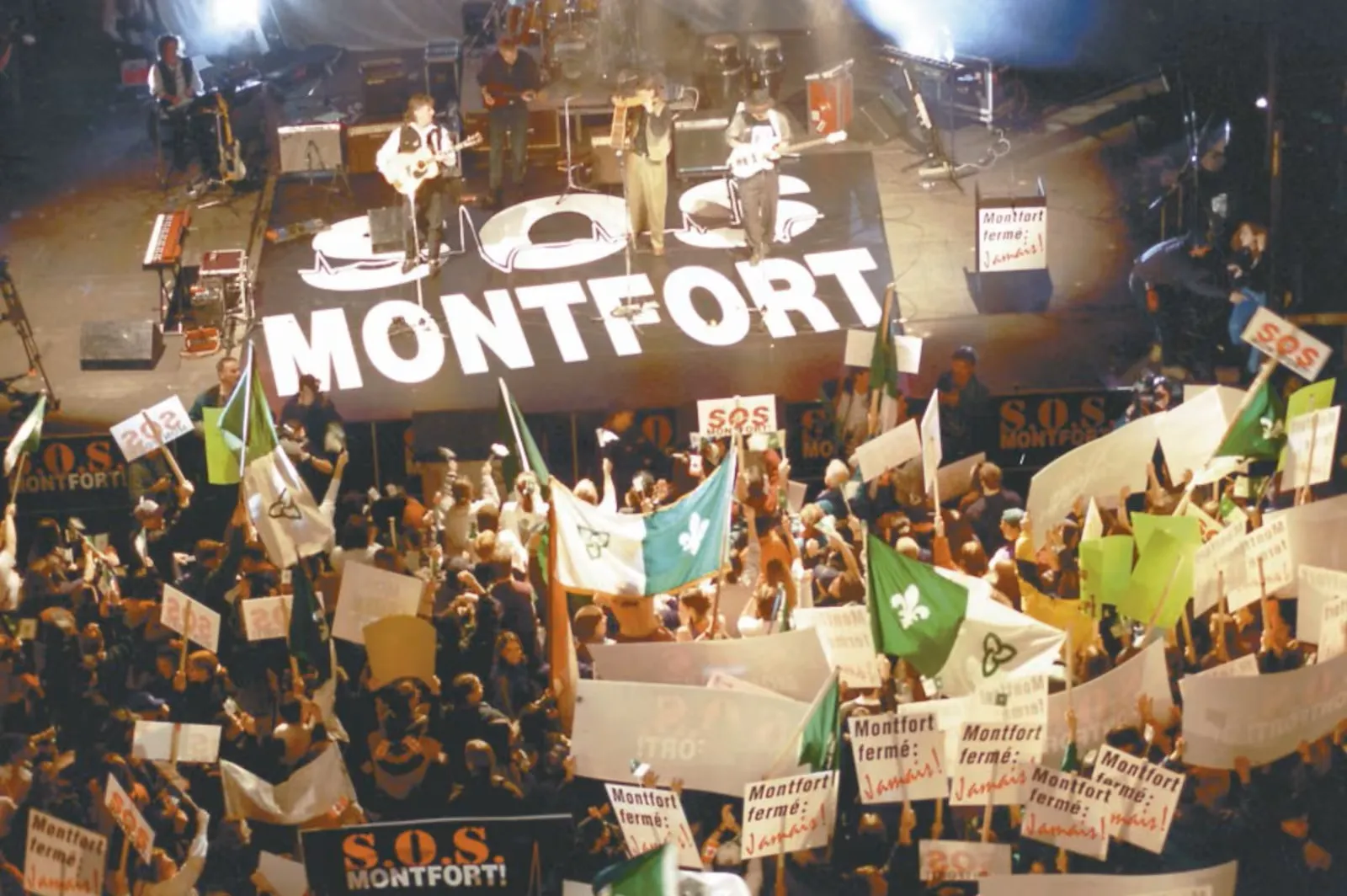 Photo montrant un groupe de musique qui joue sur une scène où il est écrit, en gros, au sol : SOS Montfort. Une foule attroupée brandit des pancartes, dont plusieurs disent : « Montfort fermé : jamais! », et des drapeaux franco-ontariens.