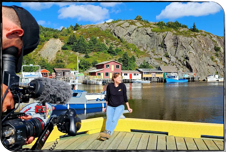 Photo de Nadia Campbell assise sur un quai dans un village côtier. À l’avant, un caméraman la filme.