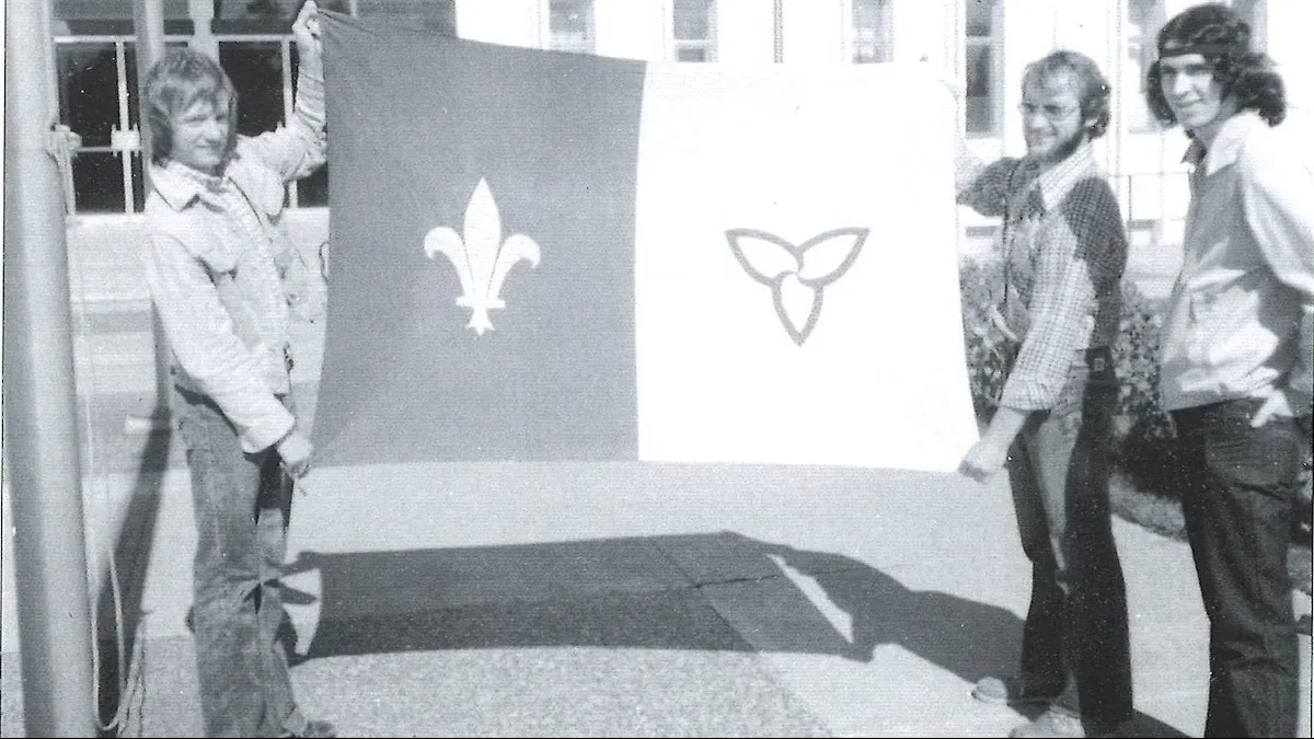 Photo en noir et blanc montrant Gaétan Gervais et Michel Dupuis tenant le premier drapeau franco-ontarien. Un autre jeune homme les accompagne.