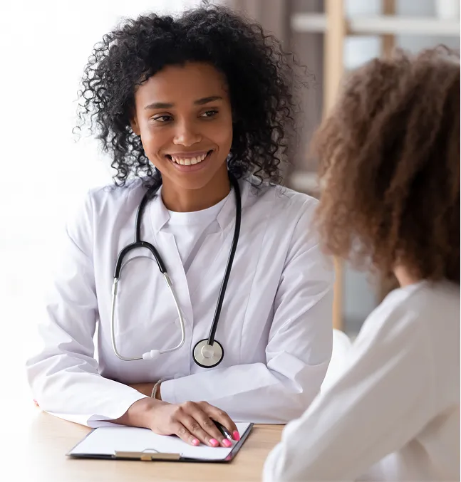 Une femme portant une blouse blanche et un stéthoscope autour du cou est assise à une table. Elle parle à une autre personne et prend des notes.