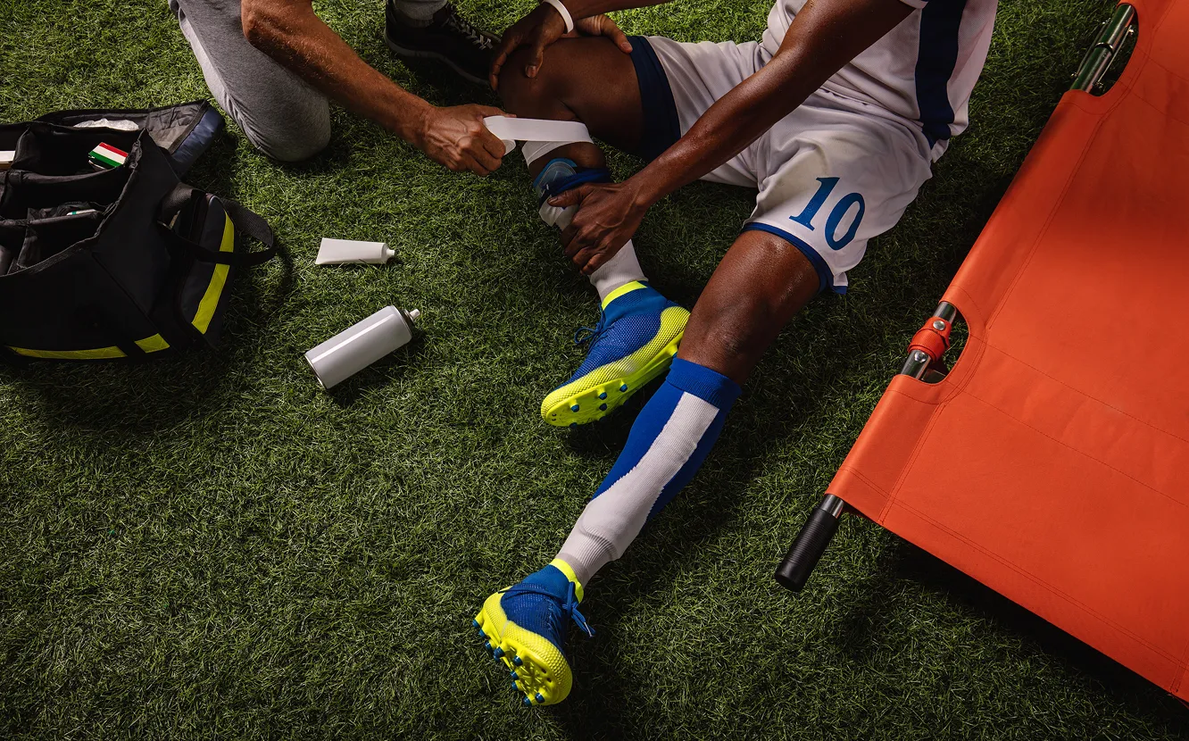 Un joueur de soccer est assis au sol. Une personne lui fait un bandage autour de sa jambe.