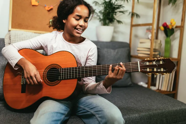 Une adolescente souriante aux cheveux frisés joue de la guitare acoustique sur un canapé.